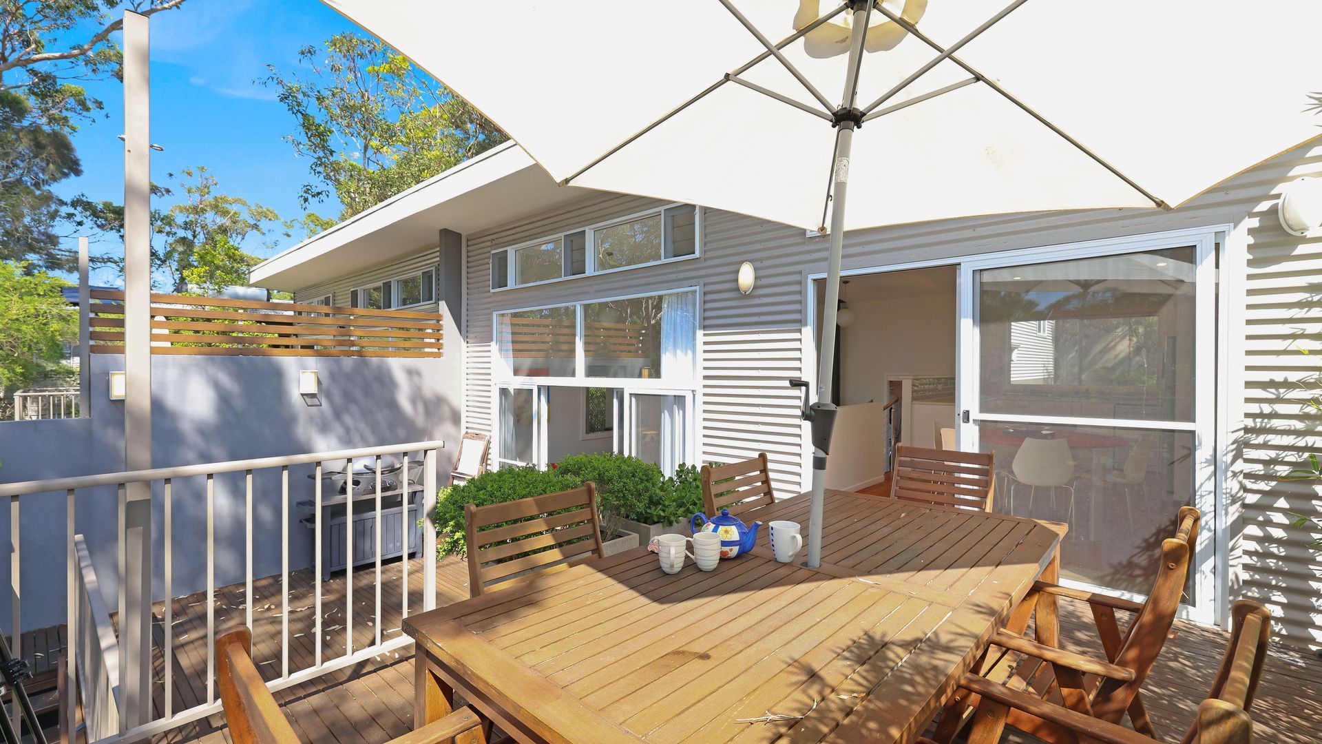 Een houten terras met een tafel, stoelen, een witte parasol en een huis met grote ramen en een balkon.
