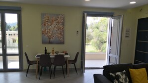 A dining area with a table set for two, a painting on the wall, and a balcony with a view of greenery. - Casa Marina apartment  kontokali  (Kontokali)