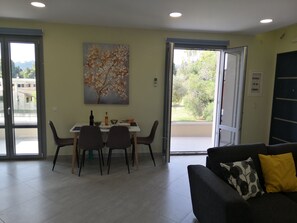 A dining area with a table set for two, a painting on the wall, and a balcony with a view of greenery.
