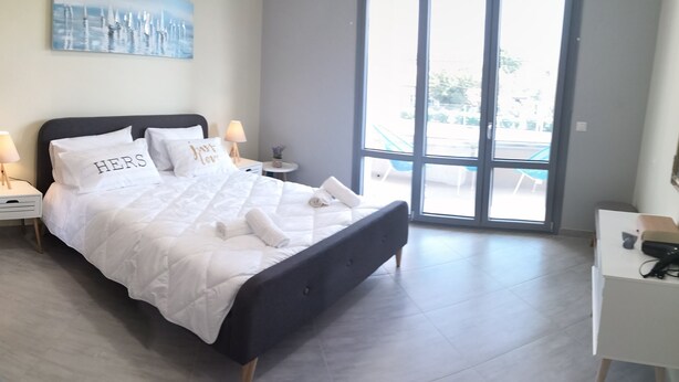 A modern bedroom with a large bed, bedside lamps, a small table, and a view through a large window. - Casa Marina apartment  kontokali  (Kontokali)