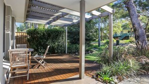 Un patio cubierto con terraza de madera, una mesa con sillas y vistas a un jardín con diversas plantas y un árbol.