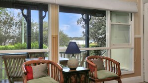 A room with a large window overlooking a beach, two wooden chairs with cushions, a small table with a lamp, and a view of a boat on the water.