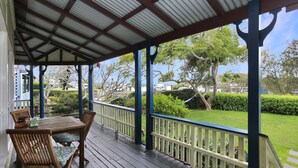 A covered outdoor area with a wooden table and chairs, a railing, and a view of a well-maintained garden and parked vehicles.