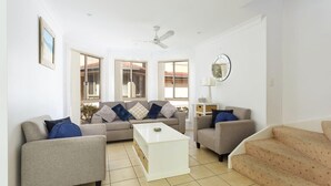 Un salon spacieux comprenant un ensemble de canapés, une table basse et un téléviseur. De grandes fenêtres laissent entrer la lumière naturelle et offrent une vue sur l’extérieur.