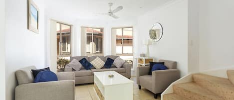 A spacious living room with a sofa set, a coffee table, and a TV. There are large windows providing natural light and a view of the outside.