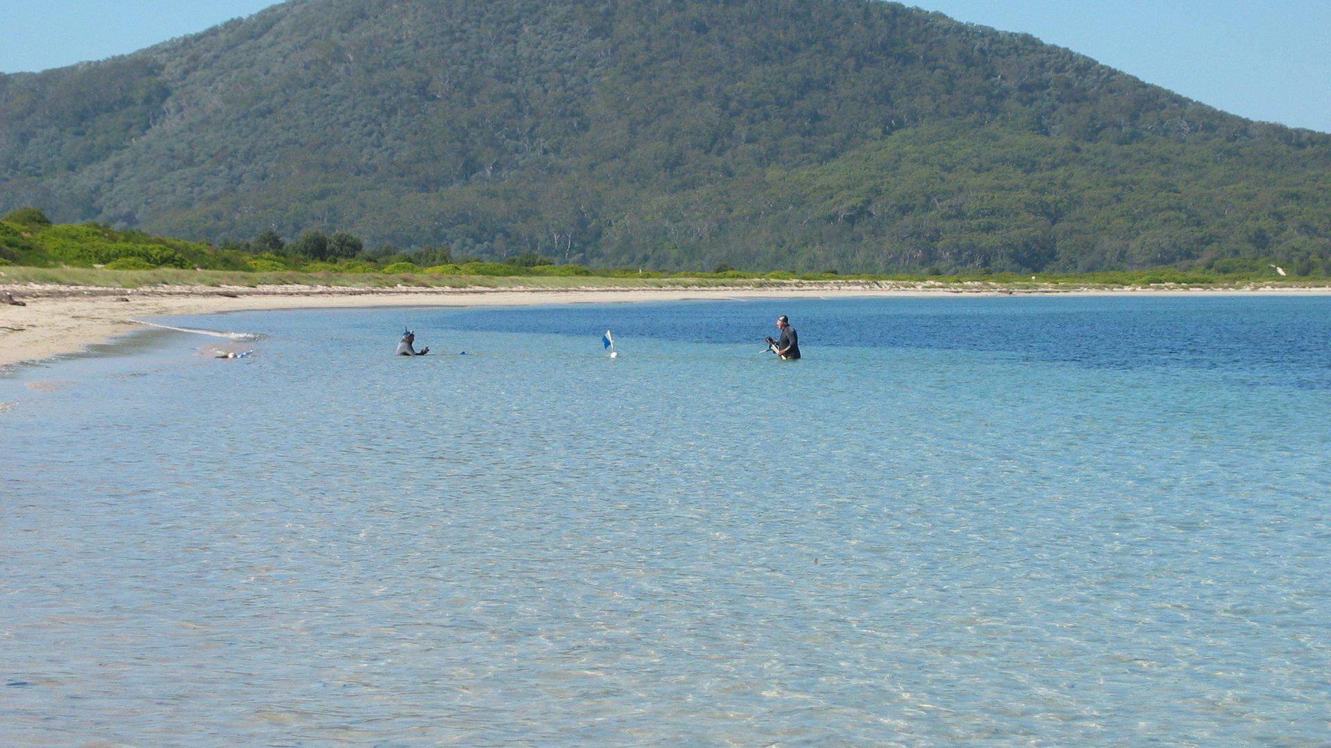 Una playa con agua cristalina, una orilla arenosa y una colina boscosa al fondo.