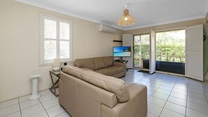 Ein Wohnzimmer mit einem beigefarbenen Sofa, einem Fernseher und einem Fenster mit Jalousien.