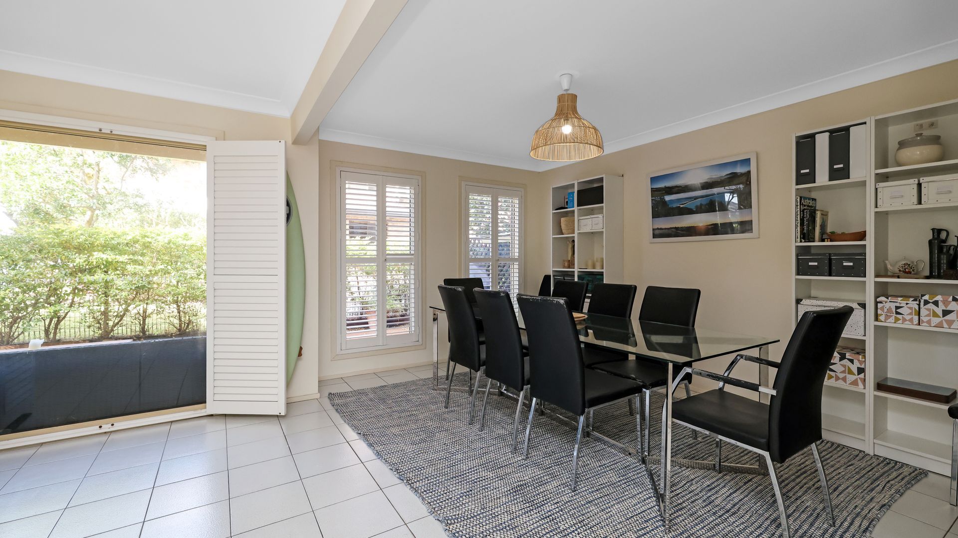 A dining area with a glass table, black chairs, a patterned rug, a ceiling light, and a view of greenery outside.