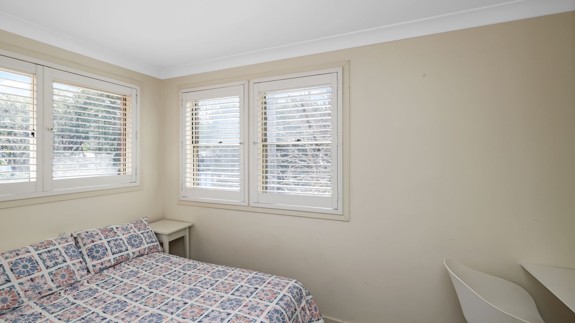 Quarto com cama, mesa de cabeceira e duas janelas com persianas.