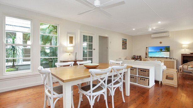 A spacious living area with a wooden dining table, white chairs, a sofa, and a television displaying a beach scene. - Beach Shack - close to the beach (Hawks Nest)