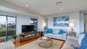 A spacious living room with a large flat-screen TV, a sofa with blue cushions, a coffee table, and a large window offering a view of the outdoors. - Myall River Magic - well-appointed townhouse overlooking the Myall River (Tea Gardens)