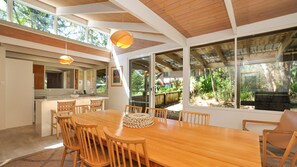 A spacious dining area with a large wooden table and chairs, a kitchen in the background, and a view of the outdoors through large windows.