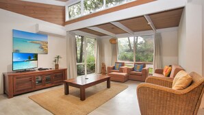 A spacious living room with a wooden ceiling, large windows offering a view of greenery, a television on a wooden cabinet, and a wicker chair. - Sandy Shores - Hawks Nest Holiday Home (Hawks Nest)