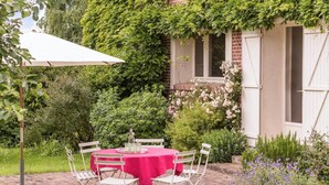 Een tuinpatio met een tafel en stoelen, omgeven door weelderige begroeiing en een wit gebouw met luiken.