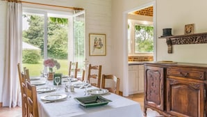 Een eethoek met een tafel voor vier personen, houten stoelen, een groene dienblad en een vaas met bloemen. Er is een raam met gordijnen, een ingelijst schilderij aan de muur en een houten kast met decoratieve bovenkant.