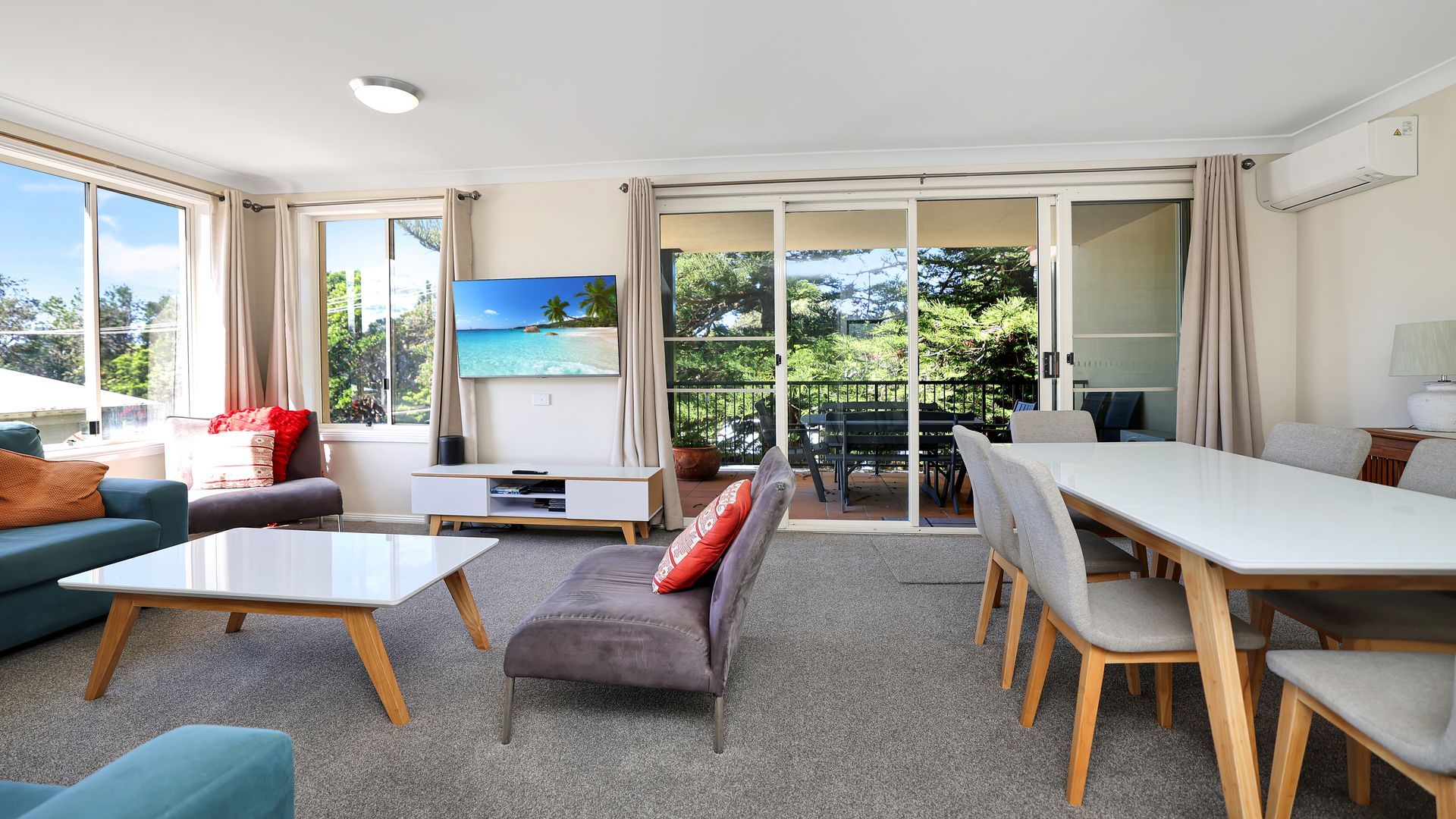 Amplia sala de estar con sofá, mesa de centro y mesa de comedor. Hay un televisor montado en la pared y un gran ventanal corredizo que da a un área exterior.