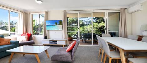 Amplia sala de estar con sofá, mesa de centro y mesa de comedor. Hay un televisor montado en la pared y un gran ventanal corredizo que da a un área exterior.