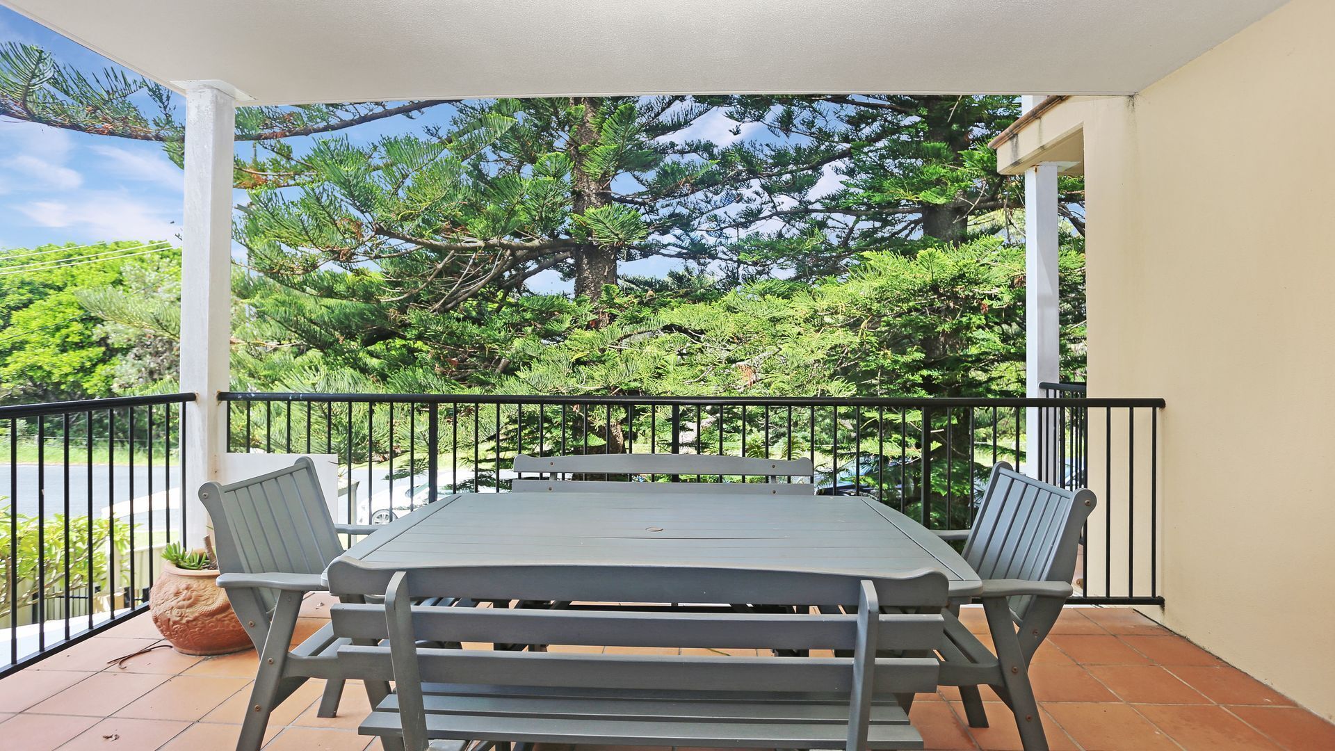 A covered patio with a table and chairs, surrounded by a black railing, overlooking a lush green landscape.