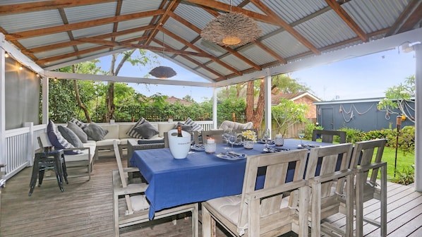 Une terrasse extérieure couverte, avec une table en bois dressée pour les repas, des sièges rembourrés et un luminaire suspendu.