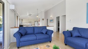 Un salon moderne avec un canapé bleu, une table basse en bois et une vue sur la cuisine et la salle à manger, grâce à une porte ouverte.