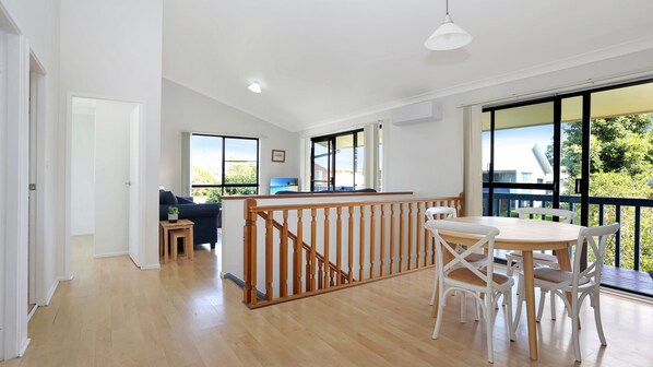 A spacious living area with a wooden staircase, a dining table with chairs, and large windows offering a view of the outdoors.