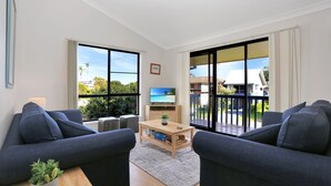 Un salon moderne avec deux canapés, une table basse et un téléviseur posé sur un meuble. De grandes fenêtres offrent une vue sur l’extérieur.