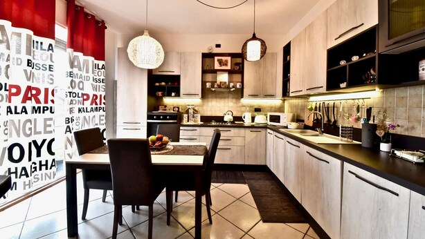 A modern kitchen with a dining area, featuring light wood cabinets, a black countertop, and hanging pendant lights. - Modern apartment between Milan and Como (Cogliate)