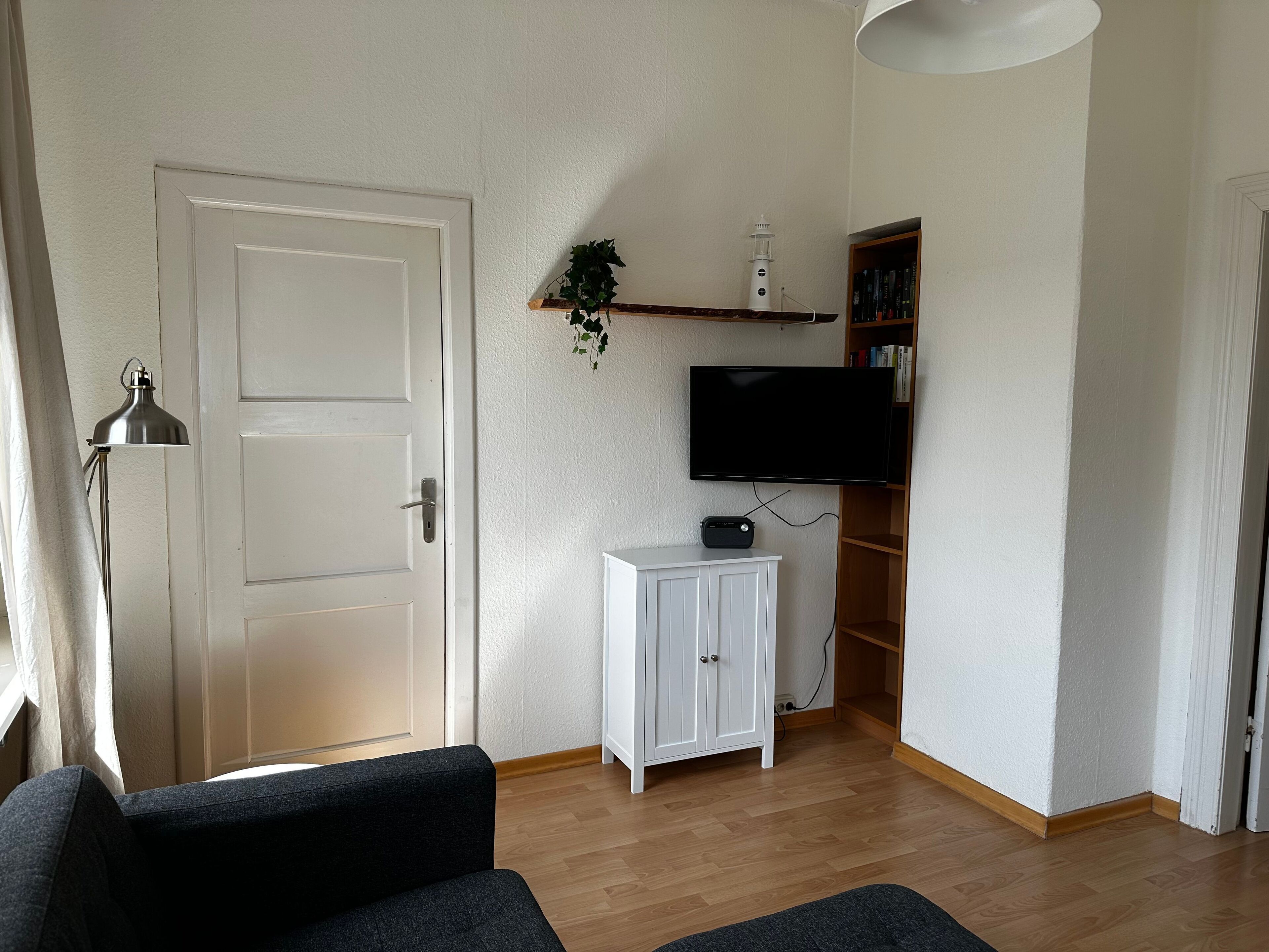 A living room with a white door, a flat-screen TV mounted on the wall, a small white cabinet, and a wooden floor.