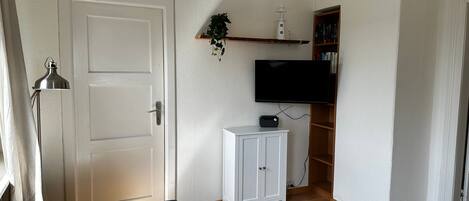 A living room with a white door, a flat-screen TV mounted on the wall, a small white cabinet, and a wooden floor.