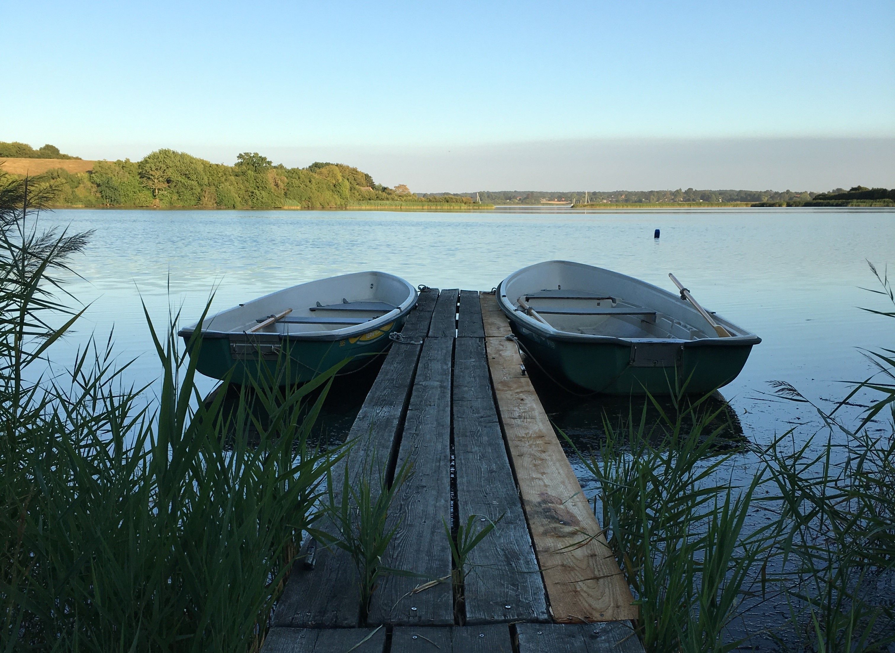 Two boats docked at a wooden pier with a calm lake and a forested hill in the background.