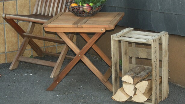 Een houten stoel met een rieten mand vol bloemen en fruit, een klein houten tafeltje en een stapelhout tegen een muur.