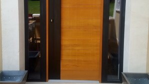 Een moderne houten deur met een zwart frame, omlijst door twee metalen plantenbakken.