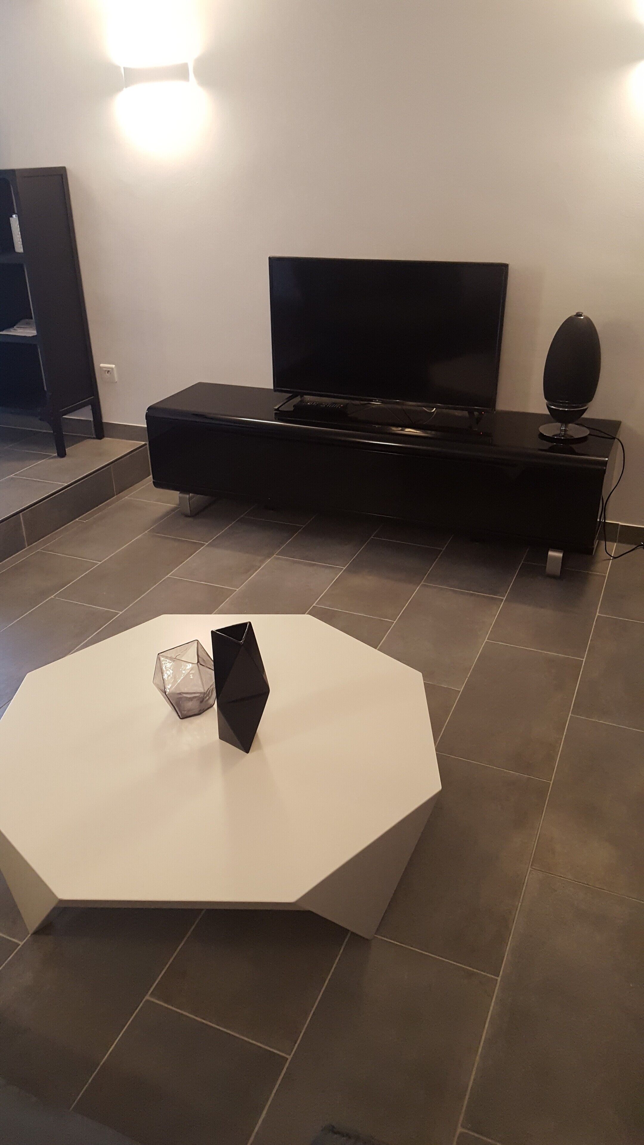 Een moderne woonkamer met een flatscreen televisie op een zwart voetstuk, een witte achthoekige salontafel en een zwarte speaker.