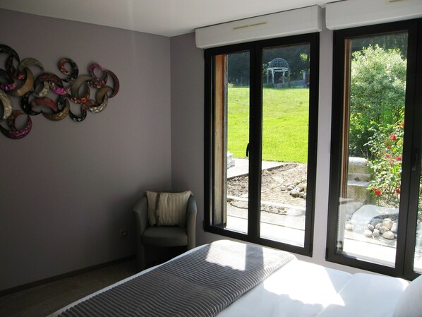 A bedroom with a bed, a chair, a wall with decorative items, and large windows with a view of a garden.