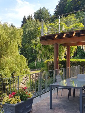 A glass table and chairs on a stone patio with a metal railing, surrounded by greenery and a wooden pergola.