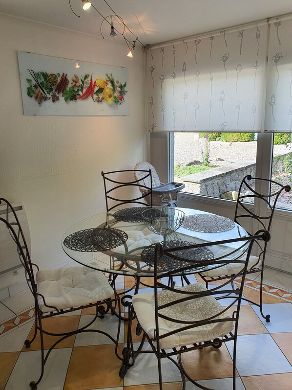 Un espace repas comprenant une table ronde en verre entourée de chaises en métal, d’un banc blanc rembourré et d’un élément décoratif mural.