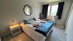 A hotel room with two beds, a bedside table with a lamp, a chair, a small table with a flower vase, and a window with curtains. - Hallingskarvet Fjellstue AS (Hol)
