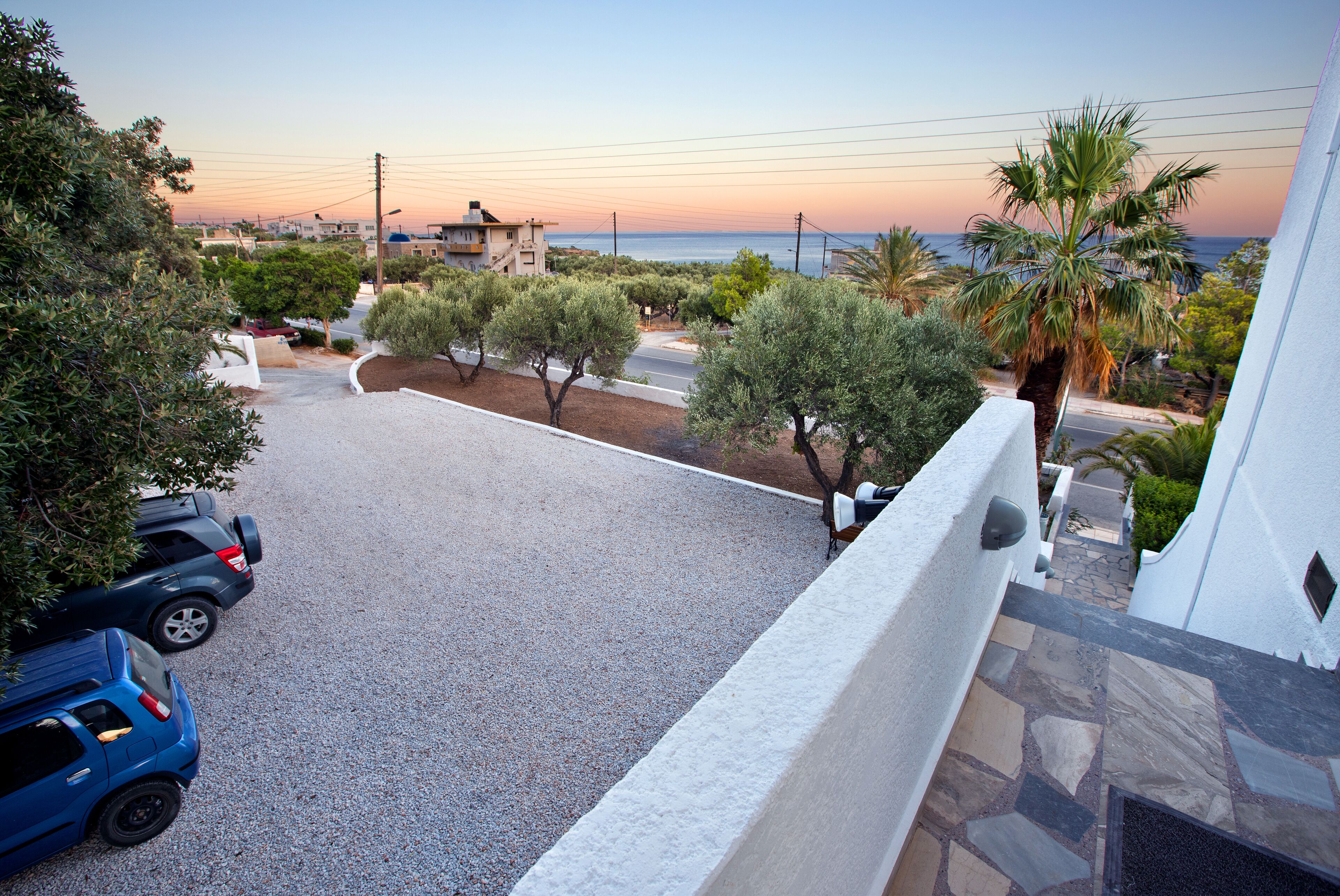 A residential area with a driveway, parked cars, and a view of the sea at sunset.