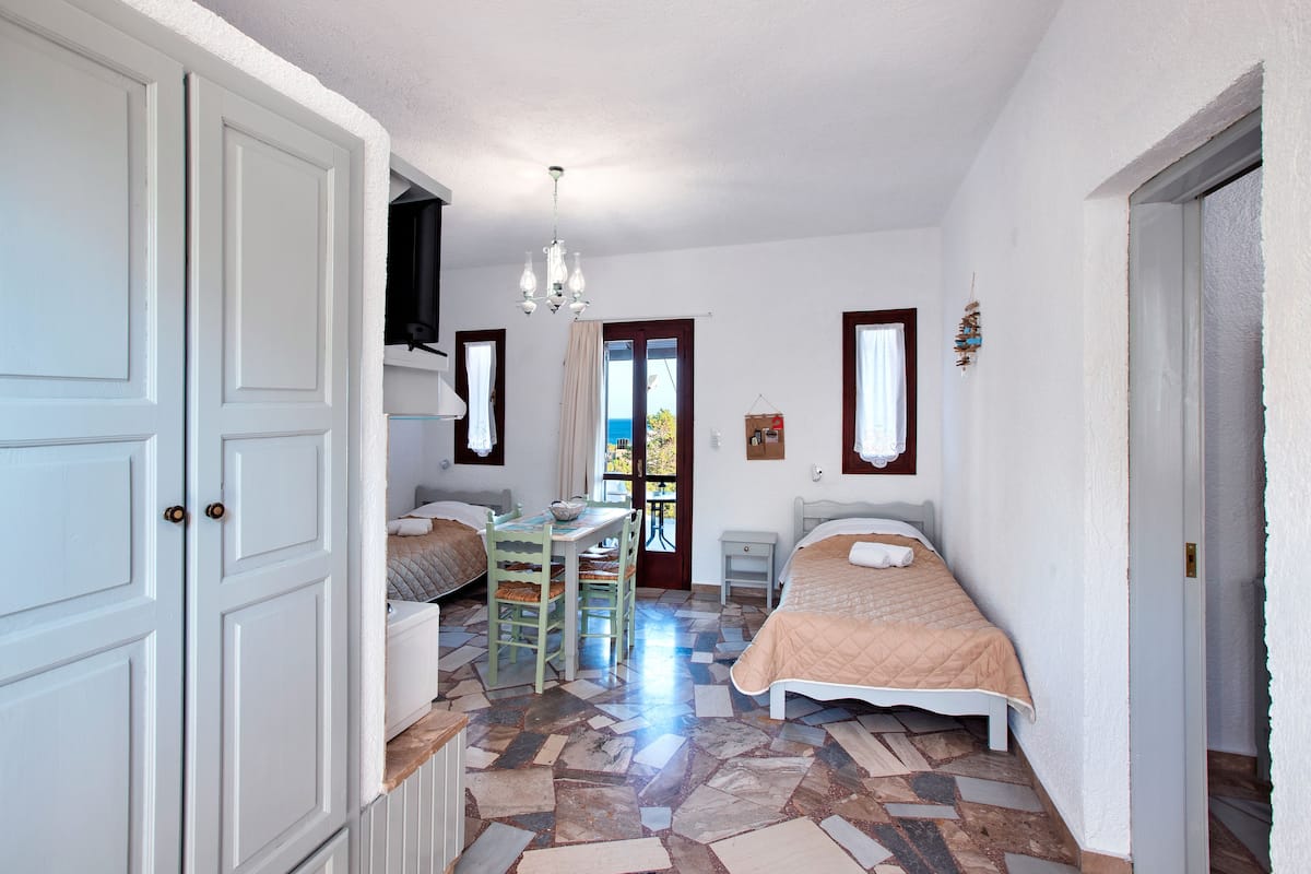 A bedroom with a bed, a table, and a chair. There is a window with curtains, a door, and a chandelier.