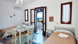 Une chambre avec deux lits, une table avec des chaises, un petit bureau et une vue sur la mer par une porte ouverte.