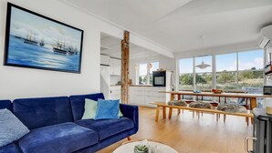 Una sala de estar moderna con un sofá azul, un comedor con una mesa de madera y una cocina con armarios blancos y un ventanal grande.