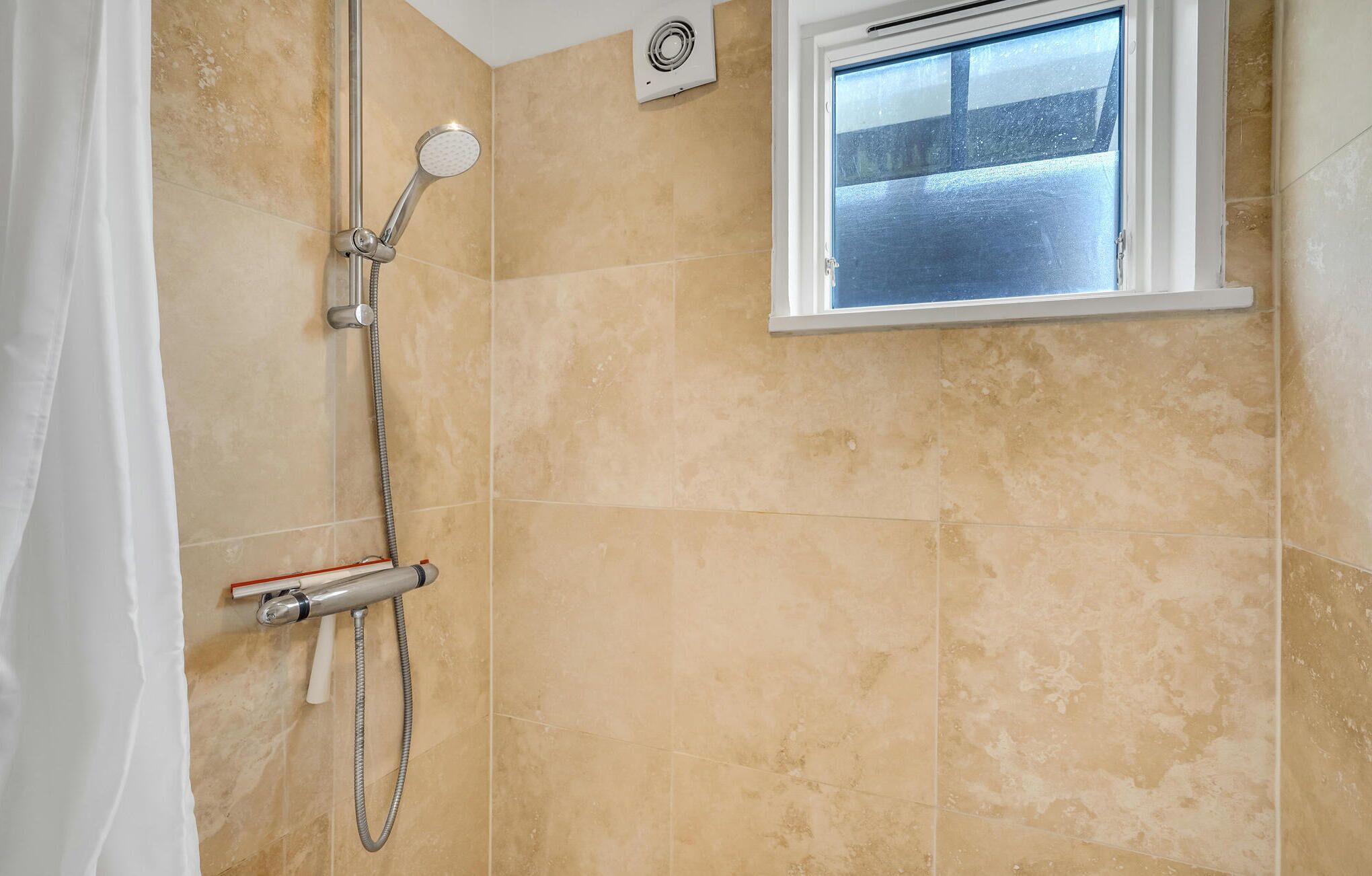 Une douche équipée d’un pommeau de douche à main et d’un robinet mural, une fenêtre avec vue et des murs recouverts de carreaux beiges.