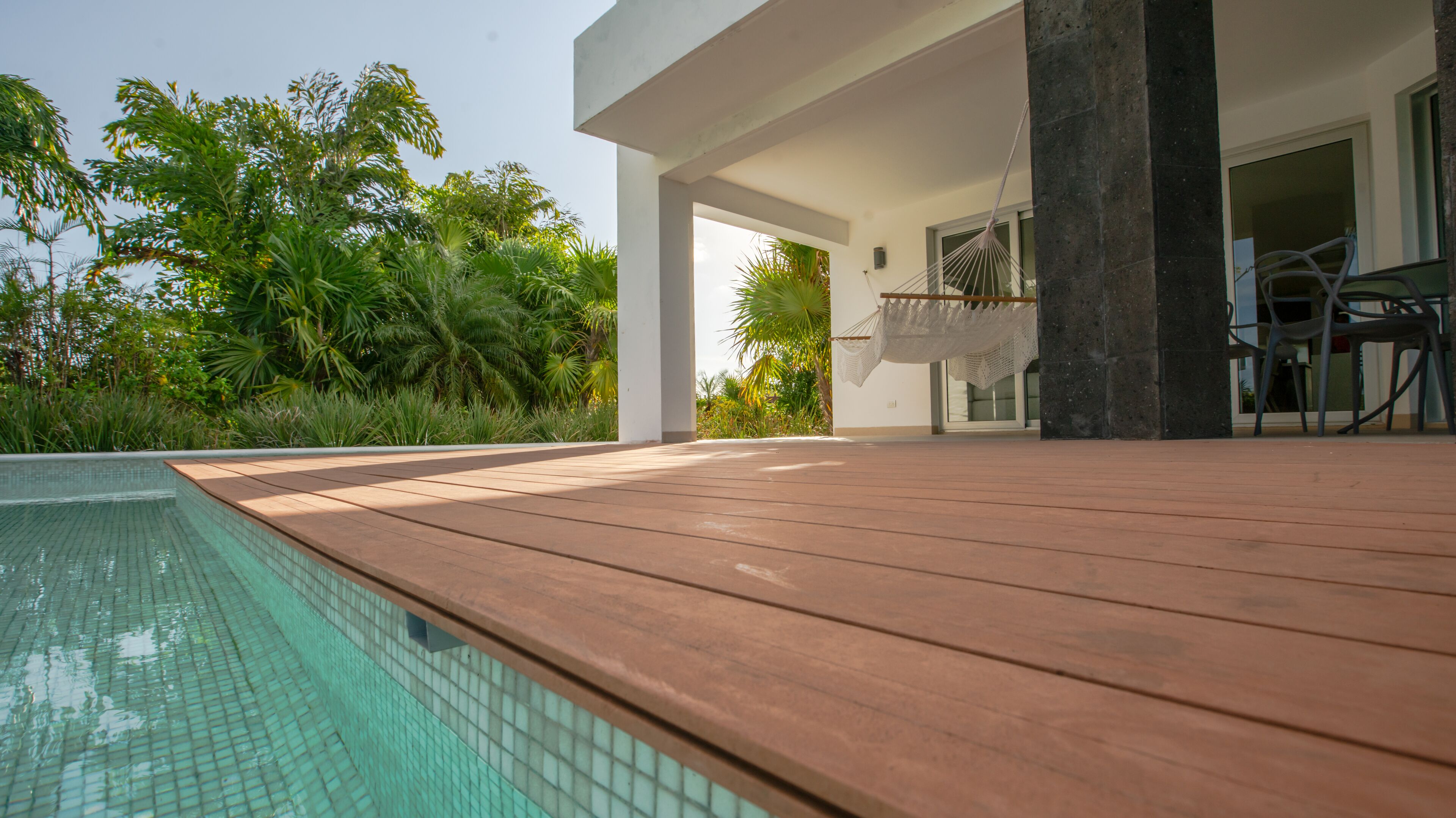A modern outdoor pool area with wooden decking, a clear pool, and lush greenery in the background.