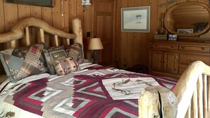 A wooden cabin bedroom with a large bed, a wooden headboard, a nightstand with a lamp, a dresser, and a mirror.