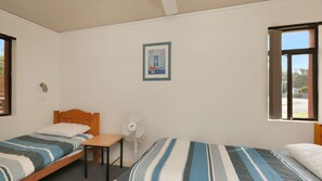 A room with two single beds, a small table, a standing fan, and a picture on the wall.