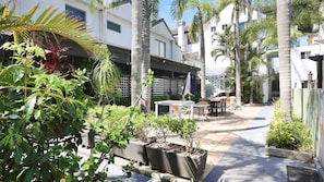 Eine Terrasse mit Sitzgelegenheiten im Freien, umgeben von tropischen Pflanzen und Palmen, die zu einem überdachten Außenbereich mit einer Treppe führt.
