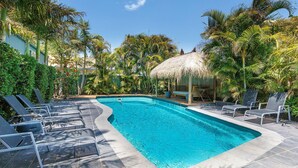 Une piscine entourée de chaises longues, bordée d’une végétation tropicale et abritée sous une structure à toit de chaume.