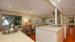 A modern kitchen with a central island, a dining area with wicker chairs, and a staircase leading to an upper level. - Patch of Paradise - Iluka (Tea Gardens)