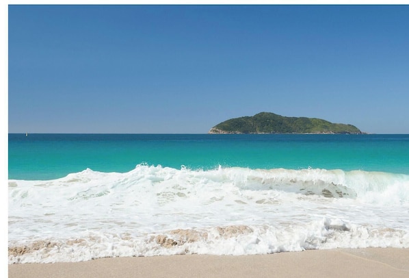 Una spiaggia con le onde che si infrangono sulla riva, un cielo azzurro limpido e una piccola isola all'orizzonte.