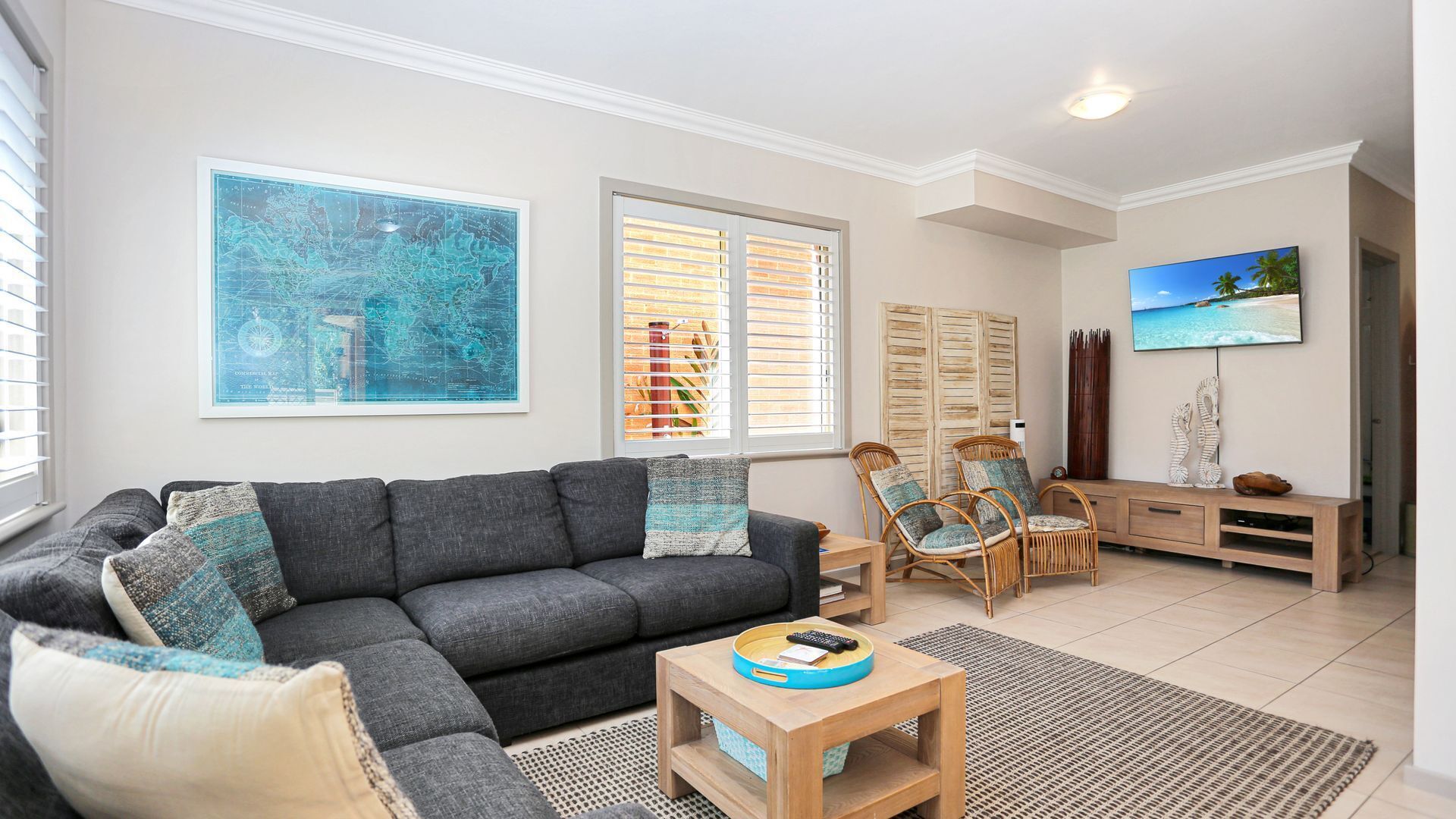 A living room with a grey sofa, two armchairs, a coffee table, and a TV on a wooden stand. There are two windows with blinds, a large abstract painting, and a wooden cabinet with decorative items.